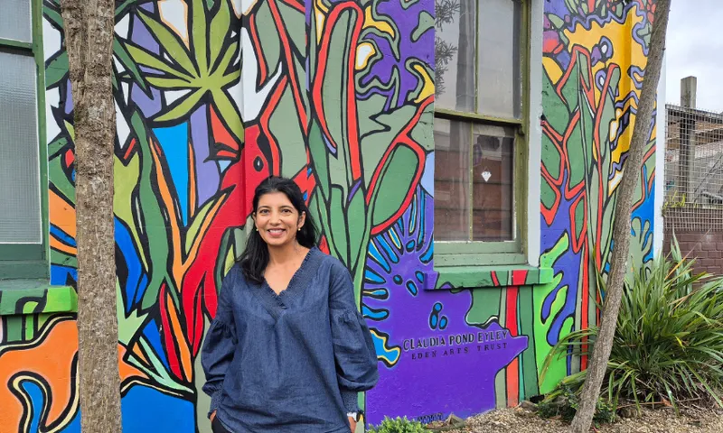 A woman with long dark hair in a blue shirt stands smiling in front of a brightly coloured mural featuring abstract plants and nature, painted on the side of a building with green-framed windows.