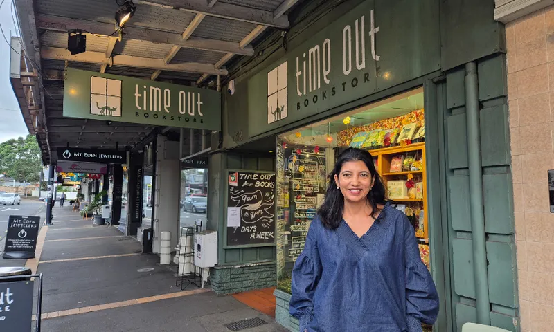 A person with long dark hair wearing a blue top stands smiling in front of the Time Out Bookstore, with colourful books visible inside and storefronts lining the street.