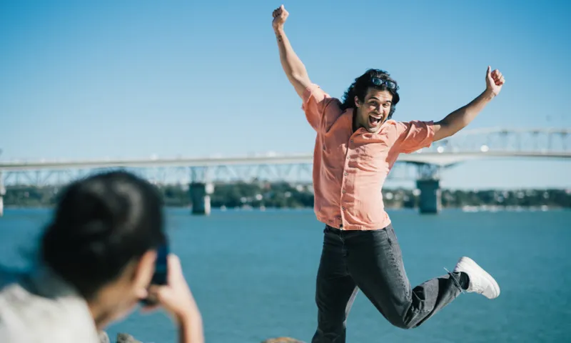 A man in a short sleeve button down shirt and jeans is jumping in the air in front of a woman who is taking a picture of him, the Auckland Harbour bridge and the sparkly, aqua harbour is behind him.