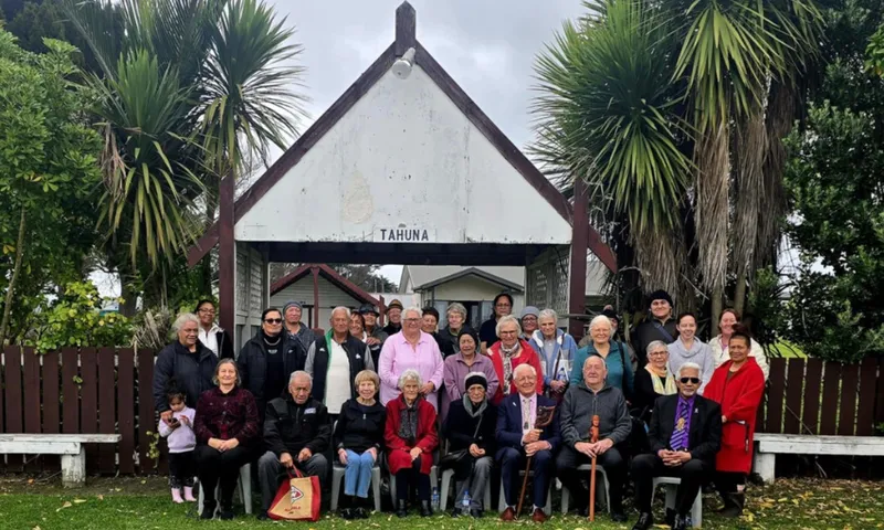 A large group of people, including elders and children, pose for a photo outside a traditional building with a sign reading "TĀHUNA." Tī kouka cabbage trees and greenery surround the scene, and everyone is dressed warmly.