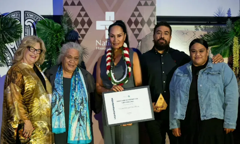 Five people stand together at an award ceremony; the central person holds a certificate and wears a floral lei. The group is dressed in formal attire, smiling in front of a decorated backdrop with patterns and greenery.