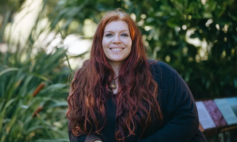 A woman with long red hair and a black top smiles while sitting outdoors, surrounded by green plants.