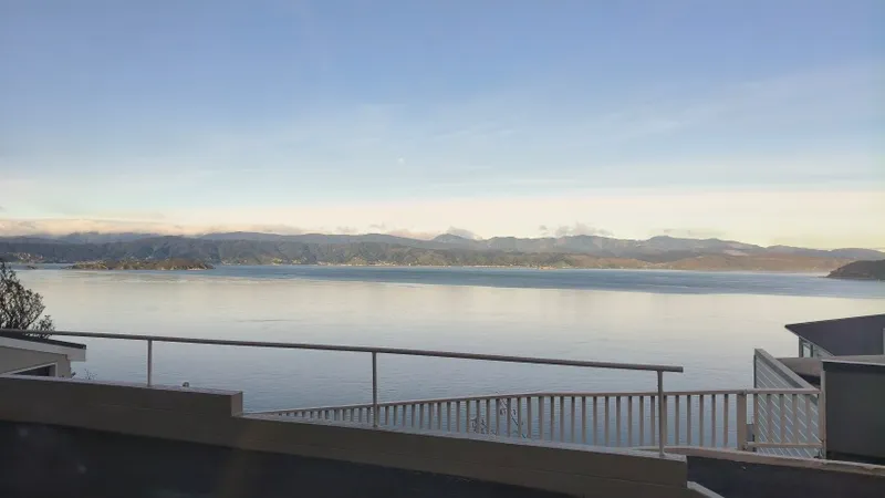 View of a calm bay with mountains in the distance, seen from behind a white railing on a balcony or terrace under a clear sky.
