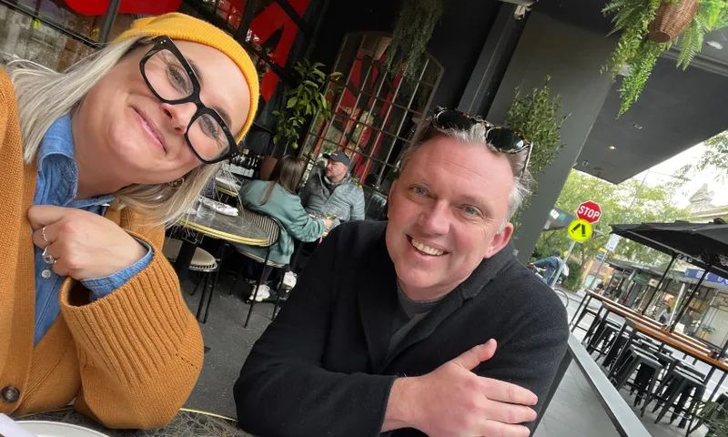 Two smiling adults sit at an outdoor cafe table. The woman wears a mustard beanie, glasses, and a tan sweater; the man wears sunglasses on his head and a dark jacket. Other people and greenery are visible in the background.