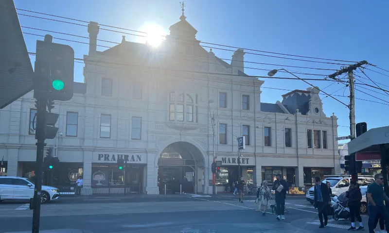People cross a busy street in front of the historic Prahran Market building in bright sunlight, with cars waiting at the intersection and the sun shining from behind the ornate facade.