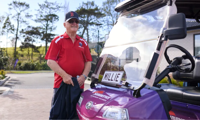 An older man in a red shirt and cap smiles while standing beside a purple golf cart labeled "MILLIE" on a sunny day, with trees and a building in the background.