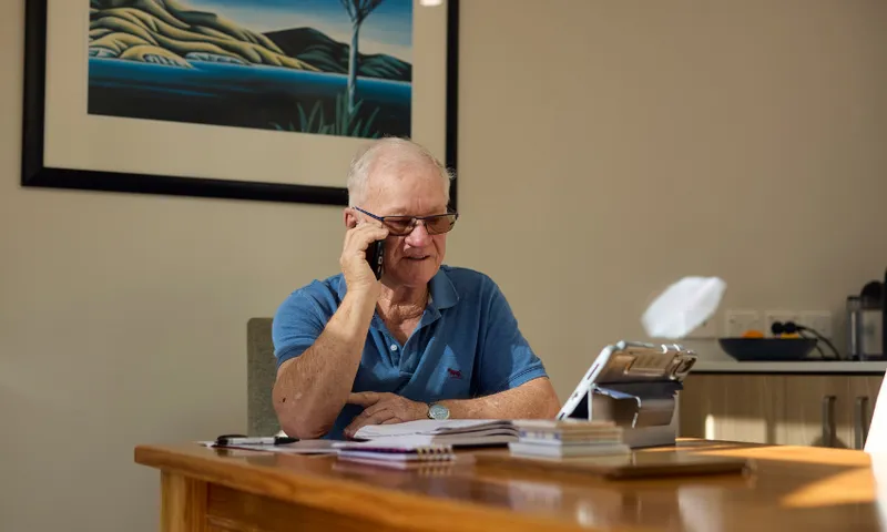 An older man wearing glasses and a blue polo shirt sits at a wooden table, talking on a smartphone. Papers, notebooks, and a tablet are in front of him. Sunlight streams in, and a landscape painting hangs on the wall behind him.