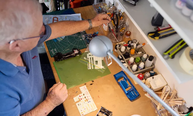 An older man works on assembling a model car at a tidy desk, surrounded by paints, tools, decals, and model car parts under a bright desk lamp.