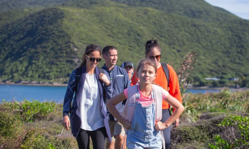 A group of five people, including a young girl in overalls, walk along a scenic outdoor trail with green hills and water in the background on a sunny day.