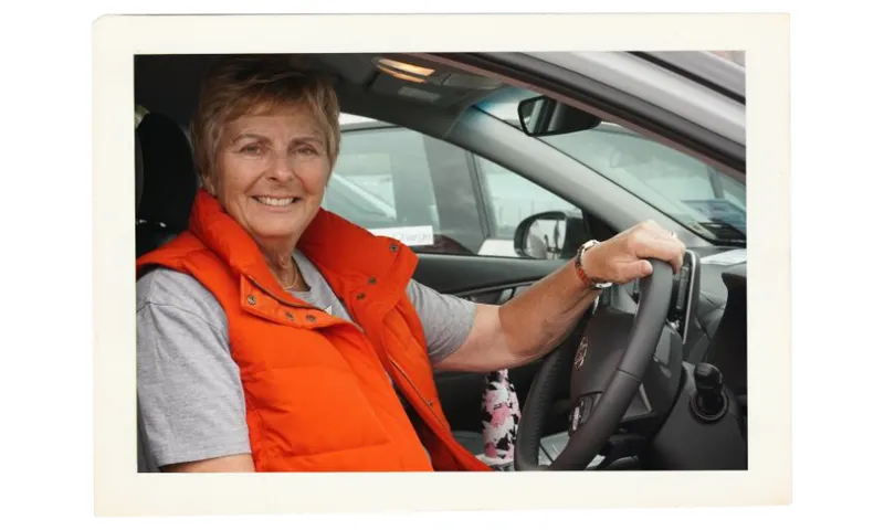 A woman wearing an orange vest and gray shirt smiles while sitting in the driver’s seat of a car, holding the steering wheel with one hand.