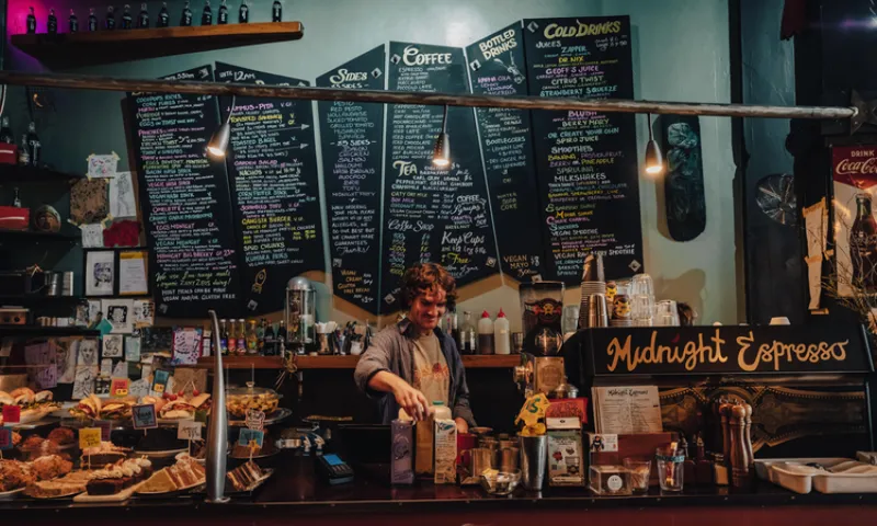 A barista stands behind the counter at a cozy café called "Midnight Espresso," with pastries on display and a large menu with colorful chalk writing hanging on the wall behind them.