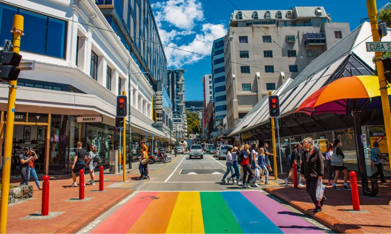A vibrant rainbow-colored crosswalk spans a busy city street lined with modern buildings. Pedestrians wait and cross under bright sunshine, with shops and a yellow-and-orange umbrella visible. Traffic lights are red.