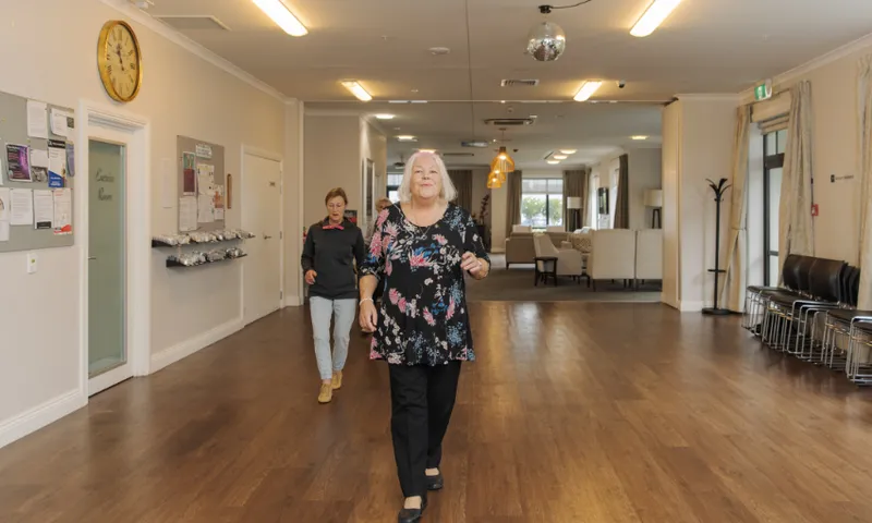 Two women walk down a spacious, well-lit hallway with wooden floors and beige walls. One woman is in the foreground, wearing a floral top, while the other follows behind. The area has chairs, a clock, and a bulletin board.