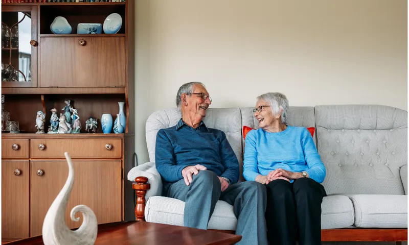 An elderly man and woman sit together on a beige sofa, smiling and laughing. Behind them is a wooden cabinet with decorative items and books, creating a cozy and warm living room atmosphere.