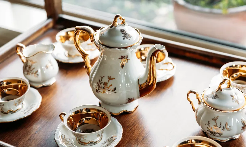 A porcelain tea set with gold accents, including a teapot, cups, saucers, a creamer, and a sugar bowl, is arranged neatly on a wooden tray near a window.