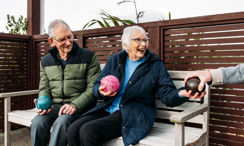 Two elderly people sit on a bench outdoors, smiling and holding colorful lawn bowls. One person hands a black bowl to the woman, who is laughing and wearing a blue quilted jacket.