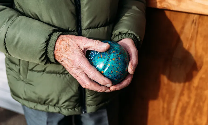 An older person wearing a green jacket holds a round, blue, speckled glass object in their hands, standing next to a wooden surface in natural light.