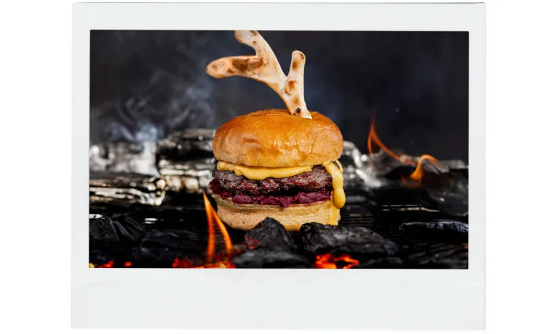 A cheeseburger with melted cheese sits on a grill above glowing coals, topped with two bread pieces shaped like antlers. Flames and smoke rise in the background, creating a dramatic effect.