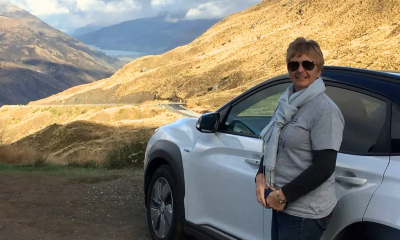 A person wearing sunglasses and a scarf stands beside a white car on a mountain road, with scenic hills and valleys in the background under a partly cloudy sky.