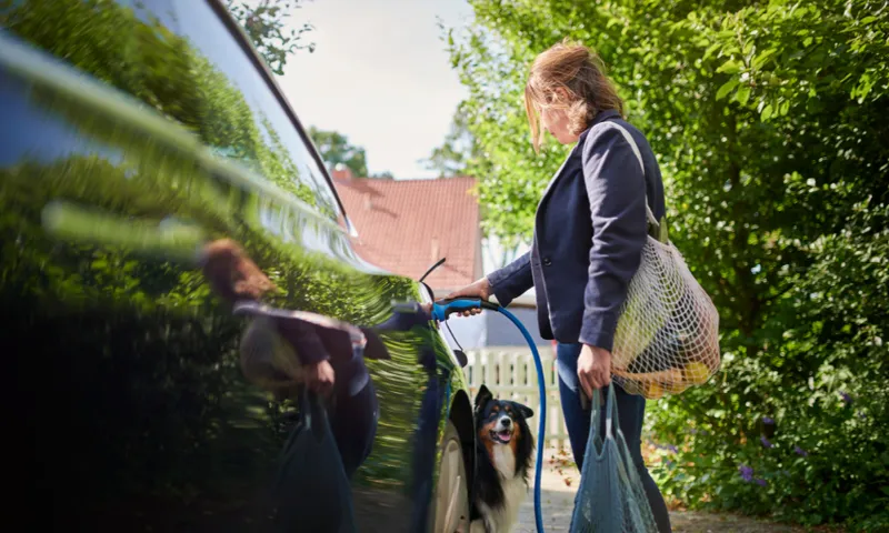 A woman stands outdoors charging an electric car, holding bags of groceries, with a black and white dog looking up at her. Trees and a house are visible in the background.