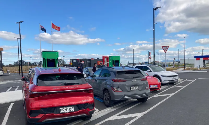 Four electric cars parked and charging at a charging station in a parking lot on a partly cloudy day, with two flags flying and several people standing near the vehicles.