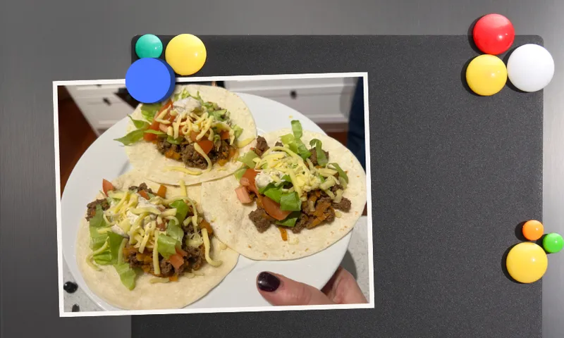 A hand holding a plate with three tacos filled with beef, lettuce, tomato, and shredded cheese is in front of a blackboard decorated with colorful magnets.