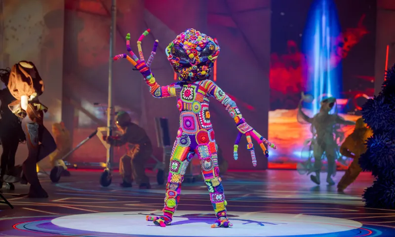 A performer in a colorful, crocheted costume with a large, multicolored headpiece and outstretched fingers stands on stage under bright lights, with people and abstract set pieces in the background.