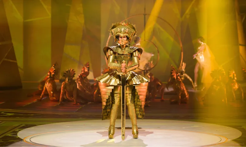 A performer in an elaborate gold costume and helmet stands center stage under dramatic yellow lighting, with dancers in gold and red outfits posing in the background.