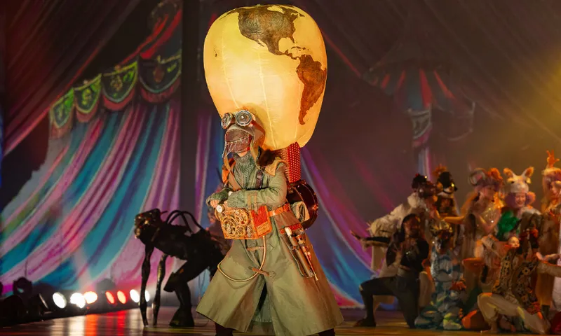 A performer in a long coat and goggles wears a large globe-shaped balloon helmet on stage, surrounded by other colorful, elaborately costumed actors under circus-like lighting and draped decorations.