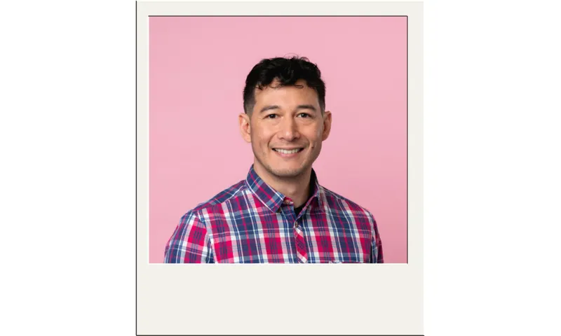 A man with short dark hair, wearing a red, blue, and white plaid shirt, smiles at the camera in front of a pink background, framed by a white Polaroid-style border.