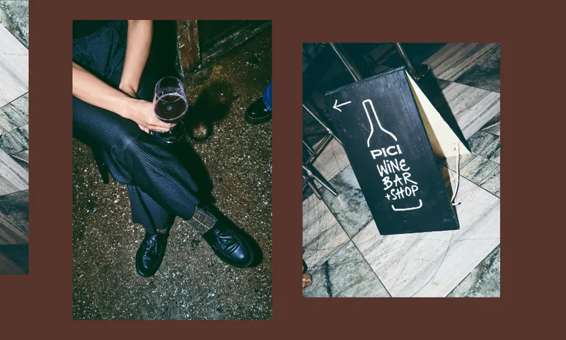 A person holding a glass of wine sits next to a chalkboard sign that reads "Pici Wine Bar + Shop" with an arrow and a wine bottle drawing, on a tiled and concrete floor.