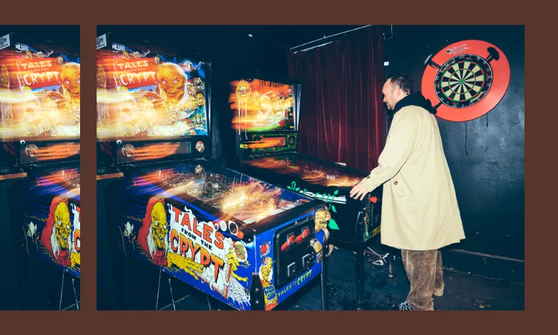 A person in a light-colored coat plays a “Tales from the Crypt” pinball machine in a dimly lit arcade. A dartboard hangs on a nearby wall above a red circle.