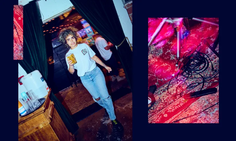 A person with curly hair, wearing a white t-shirt and ripped jeans, holds a drink while walking through dark green curtains. Beside them, a photo shows tangled cables on a red patterned rug.