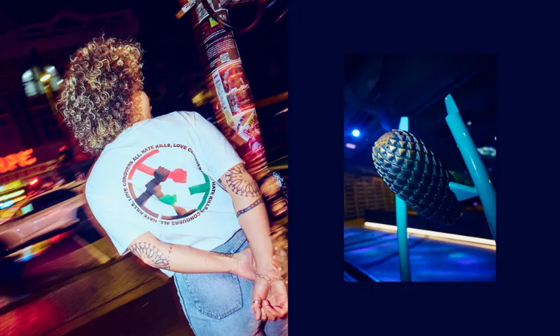 A person with curly hair stands on a city street at night, hands behind their back, wearing a white t-shirt with a colorful circular design. Beside them is a close-up of a black spiked microphone under blue lighting.