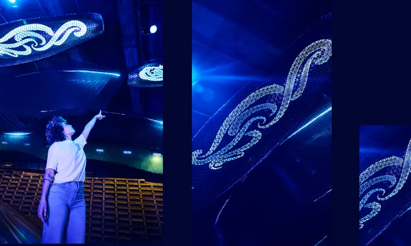 A person standing under large, illuminated, wave-shaped sculptures with intricate patterns, reaching upward in a space lit with vibrant blue lighting. Rows of empty seats are visible in the background.