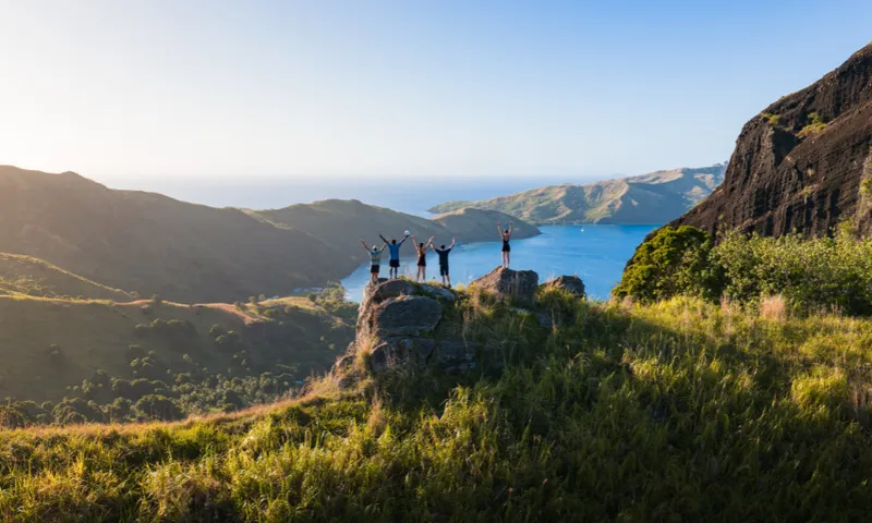 A group of people stand on a rocky outcrop, raising their arms in celebration, overlooking a scenic landscape of green hills, blue sea, and distant islands under a clear sky.