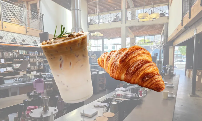 A photo of a modern coffee shop interior with an iced coffee and a croissant digitally superimposed in the foreground. The shop has high ceilings, large windows, and industrial decor.