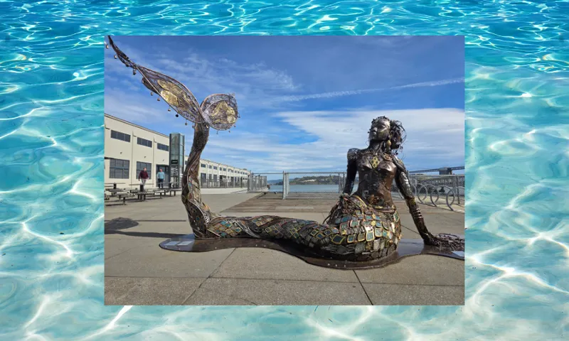 A bronze mermaid sculpture with mosaic details sits on a waterfront promenade under a blue sky, with industrial buildings and benches in the background, overlaid on an image of clear blue water.