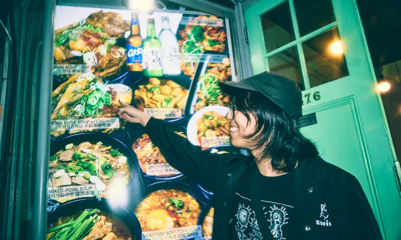 A person in a black jacket and cap excitedly points at a brightly lit restaurant menu display featuring various dishes, bottles, and colorful food photos near a door marked with the number 76.