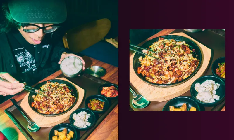 A person wearing glasses and a cap prepares to eat a sizzling dish with chopsticks, surrounded by small bowls of side dishes including dumplings and kimchi, seated at a wooden table.