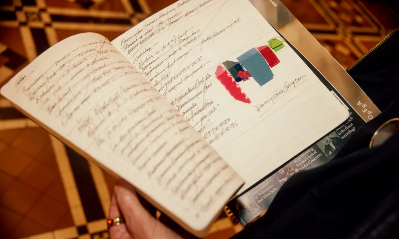 A hand holds an open notebook with handwritten text and a colorful abstract shape sketched on one page; the background features a patterned floor.