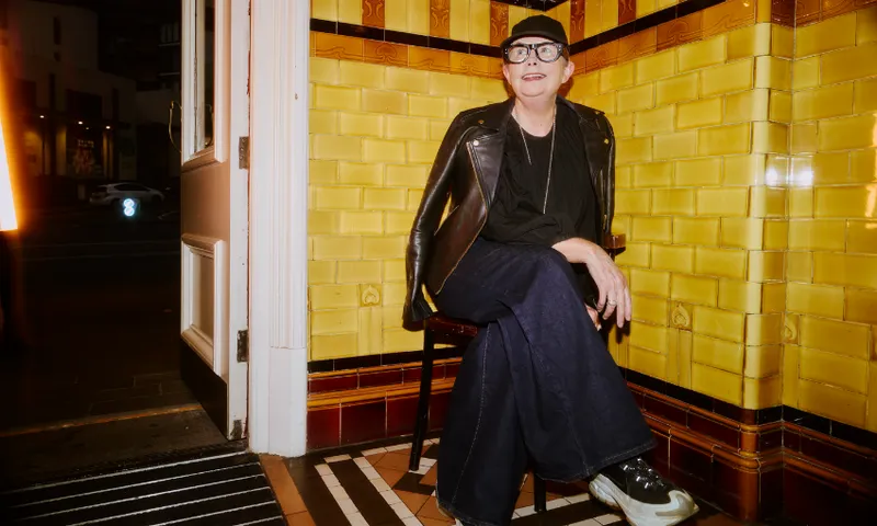 A person wearing a black cap, glasses, black leather jacket, and wide-leg jeans sits on a chair by an open door in a room with yellow tiled walls and patterned floors. It is evening outside.