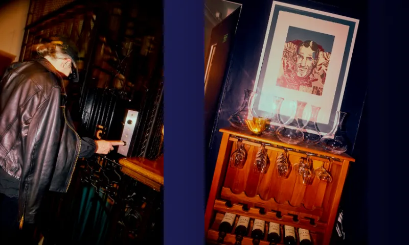 A person wearing a leather jacket presses an old-fashioned elevator button. Next to them is a wooden wine rack with bottles, decanters, and a framed photo of a smiling man displayed above.