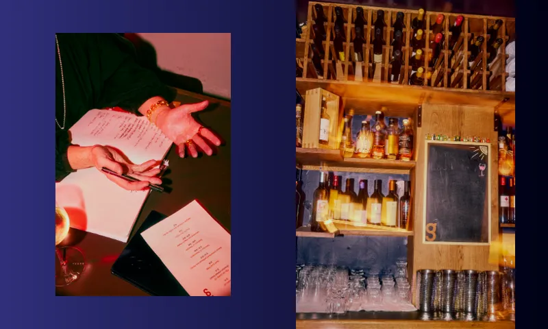 A person gestures over a menu and notebook with a glass of wine nearby; next to them, a bar shelf displays bottles, wine glasses, and a chalkboard.
