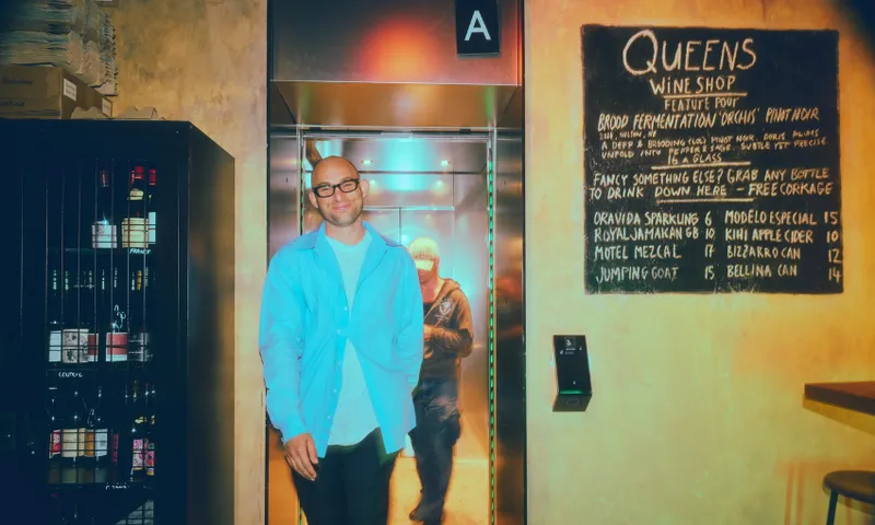 A smiling man in glasses and a light blue shirt stands by an elevator. Next to him is a chalkboard menu for a wine shop and shelves with bottles. The background is warmly lit, giving the setting a cozy atmosphere.