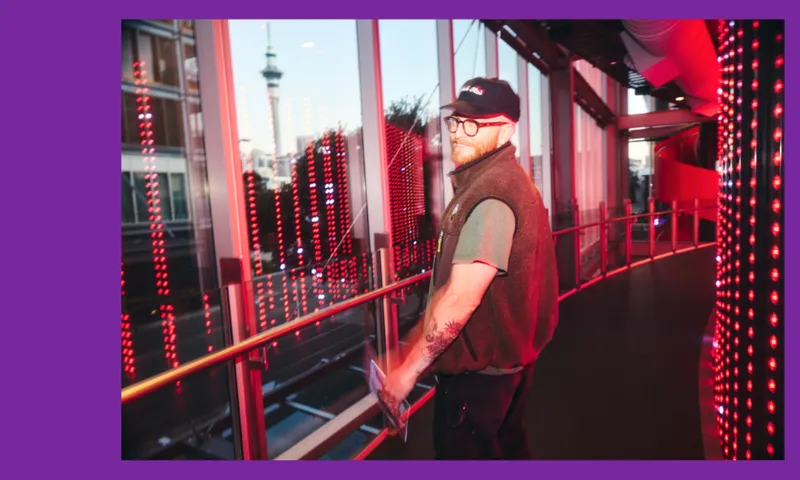 A person with glasses and a cap stands indoors by large windows, bathed in red and pink lights. City buildings and a tall tower are visible through the glass. The scene has a modern, urban atmosphere.