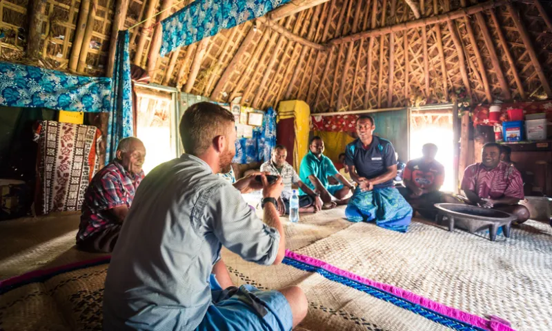 five fijian men and one white man sit on woven mats in a wooden structure. the white man holds a coconut shell filled with kava