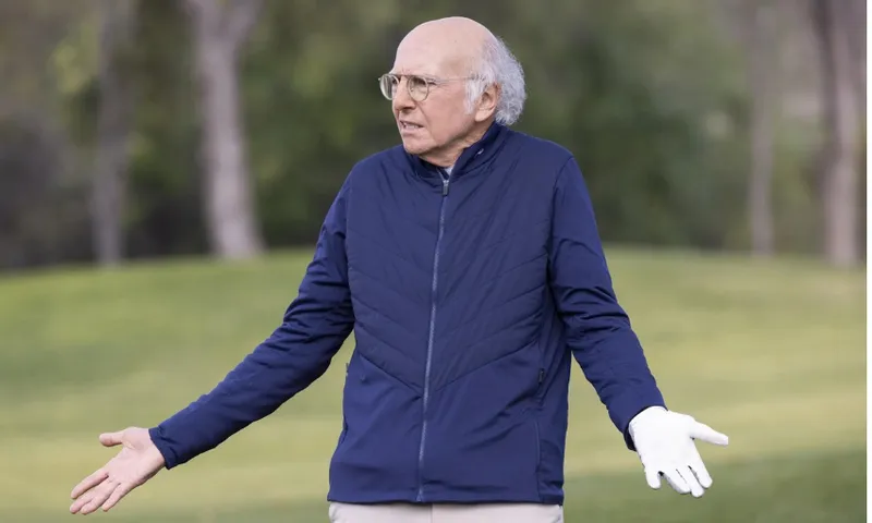 older balding white guy with white hair wears a thin black windbreaker and golfing gloves, shrugging on a golf course