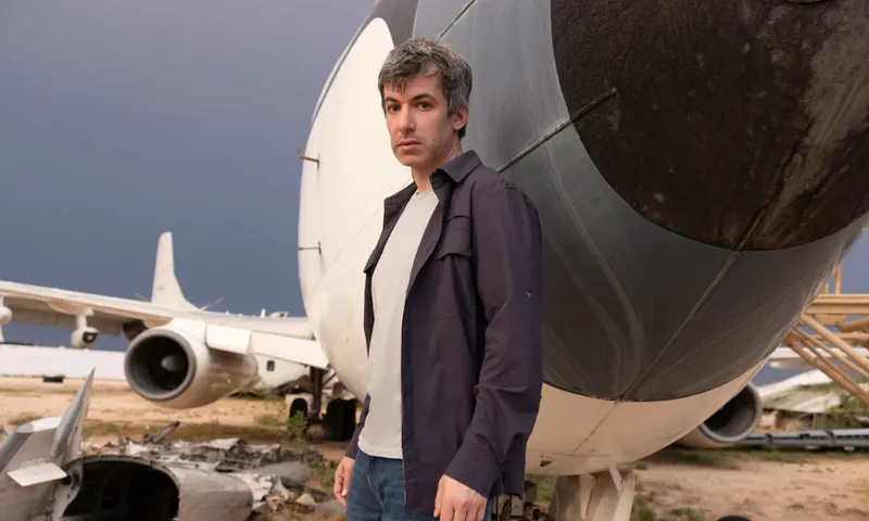 a middle aged white man with salt and pepper short hair stands in front of a plane wearing a blue shirt, white t-shirt and jeans