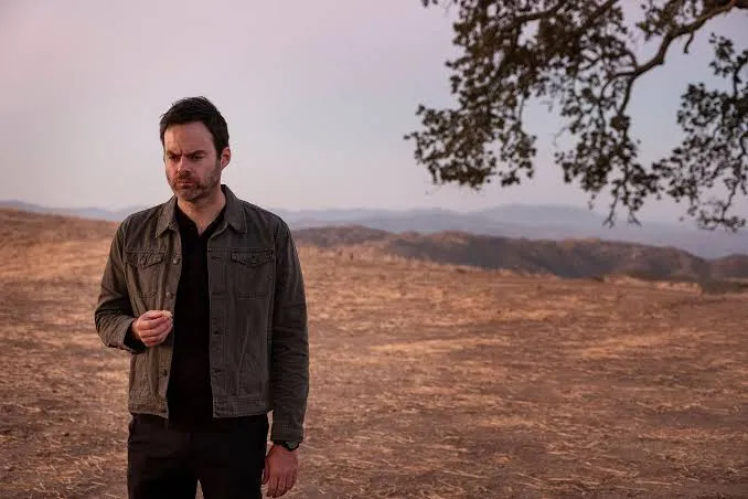 a middle aged white man with short brown hair and a beard stands on a barren hill wearing a khaki overshirt and dark jeans